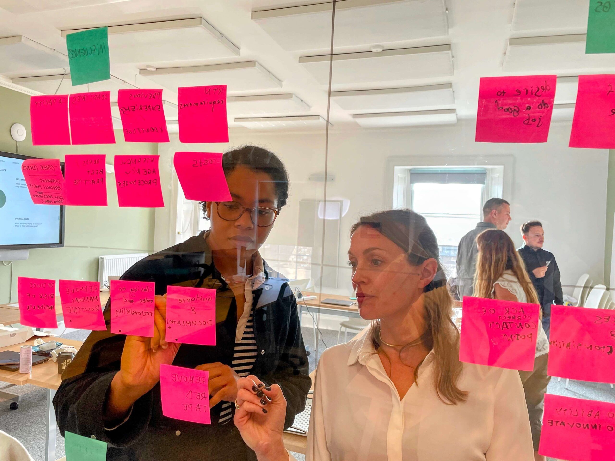 A close-up of two women collaborating in a workshop. They are standing in front of a glass wall covered in pink sticky notes, brainstorming ideas. The woman on the right, with blonde hair and a white shirt, holds a black marker and looks intently at the notes. The woman on the left, wearing glasses and a black jacket, points to one of the sticky notes. In the background, other participants are visible in the bright, modern office space.
