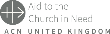 The logo for Aid to the Church in Need United Kingdom. On the left sits a dark gray circle containing a white, stylized linear symbol that resembles a cross transforming into an arrow pointing right. Beside it, the words 