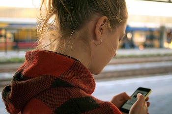 A person with light brown hair pulled back, wearing a red and black plaid hooded jacket, is looking down at a smartphone they are holding in their hands. Only the back of their head and shoulder are visible, and they appear to be outdoors or in a public space, with a blurred background that suggests a train station or similar setting.