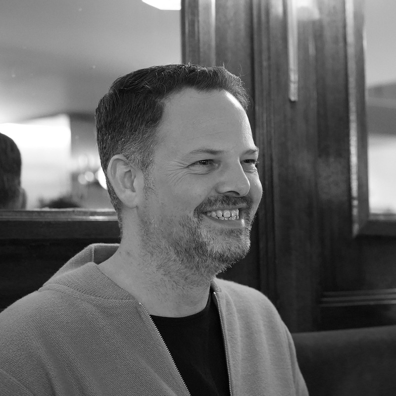 Black and white image of a man (Anthony) sat in a bar, smiling.