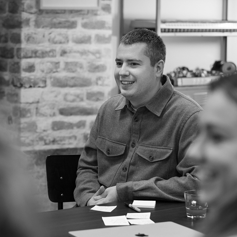 A man wearing a heavy grey shirt, sat in a workshop, smiling.