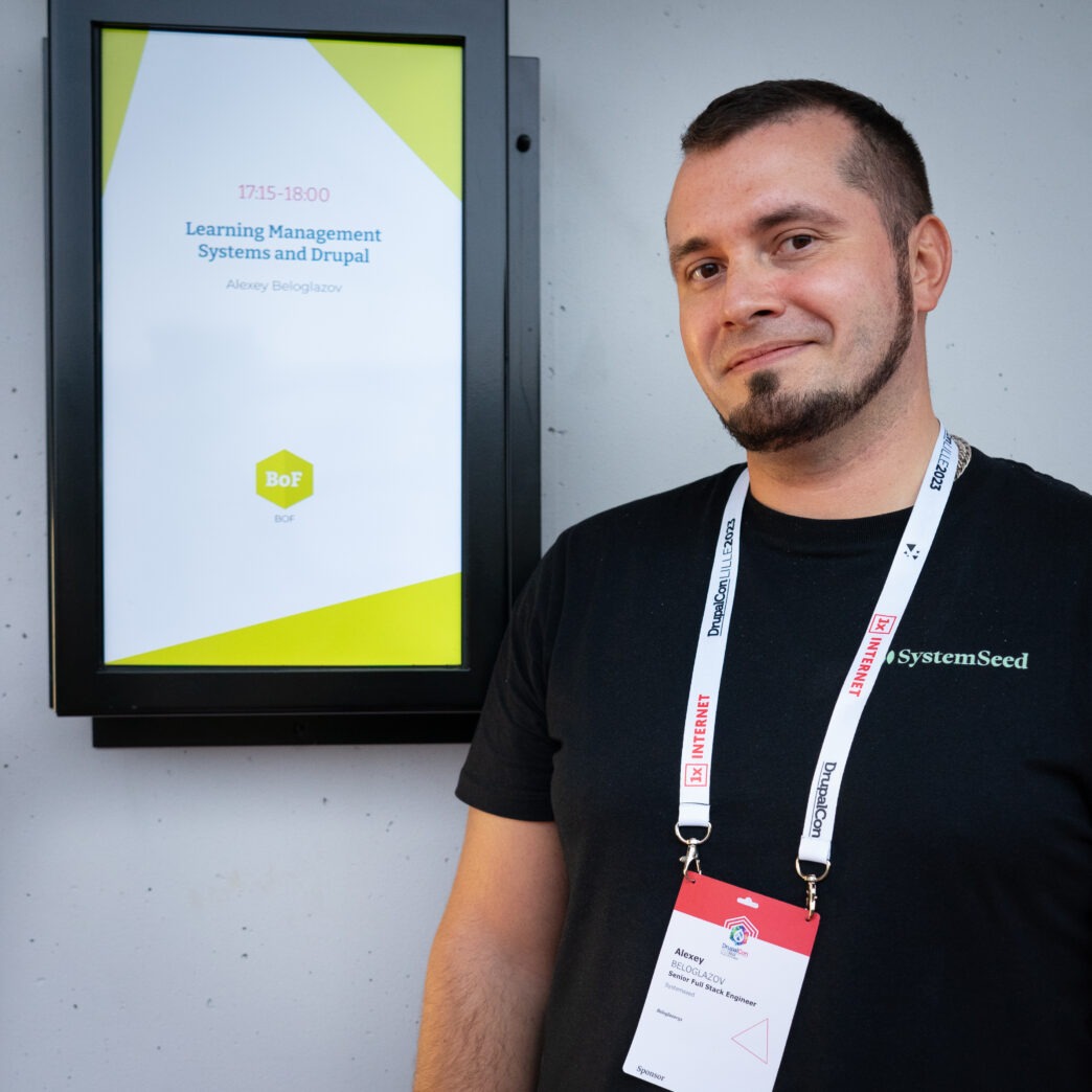 A man with short dark hair and a goatee, wearing a black t-shirt and several lanyards with ID badges, stands in front of a digital display. The display shows a presentation title "Learning Management Systems and Drupal" and the speaker's name "Alexey Beloglazov". The man is smiling slightly and looking towards the viewer.