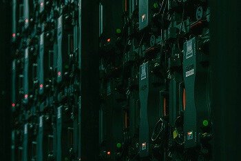 A dimly lit, close-up shot of the back of a large LED video wall or display. The wall is composed of numerous rectangular modules lined up vertically, each secured by mounting hardware. Visible on the back of the modules are power supplies/receiving cards with cables and connectors plugged into them. Some of these devices have small indicator lights (likely red and green) visible. The overall mood is dark and technical, suggesting the sustainable power source is at work, perhaps in a data center or control room environment.