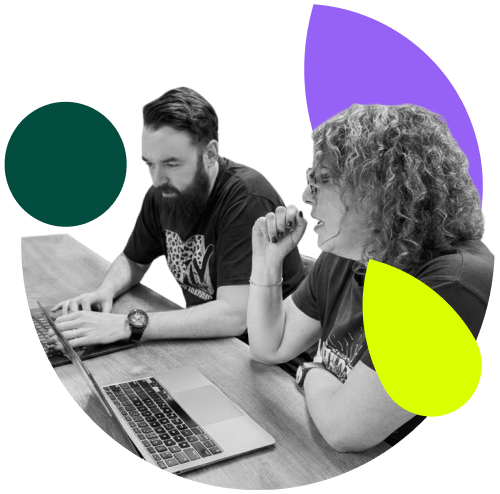 A man and a woman sit at a wooden table with laptops. The image is mostly grayscale, except for a few abstract, colored shapes around the edges. A dark green circle is in the upper left, a purple and white abstract shape is in the upper right, and a neon yellow tear-drop shape is in the lower right. The man on the left has a beard and is looking down at his laptop, typing. The woman on the right has curly hair and is looking towards the man, talking with her hand near her mouth. Both are wearing t-shirts.