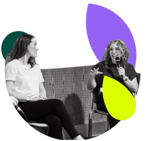 Two women are seated next to each other, as if on a panel or in an interview. The woman on the right has curly hair and glasses, and is speaking into a microphone while gesturing. The woman on the left is listening to her. The photo is in black and white and cropped into an abstract shape, with added graphic elements in dark green, purple, and neon yellow.