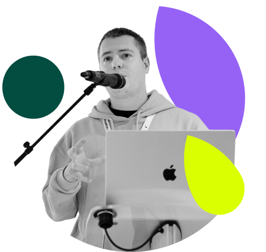 A greyscale image shows a man speaking into a microphone with an Apple laptop in front of him. He is wearing a hoodie. Abstract shapes in purple, green, and yellow are behind him.
