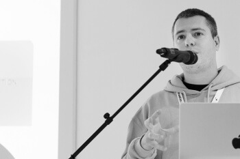 Black and white photo of a presenter speaking into a microphone on a stand, gesturing with their hand, with a laptop visible on the desk.