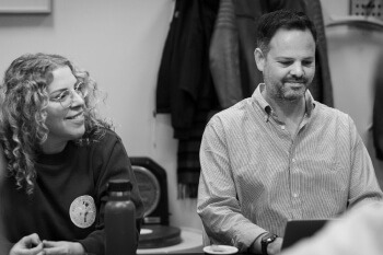 Black and white image of a man and a woman sitting at a table, smiling and engaged in conversation or a meeting.