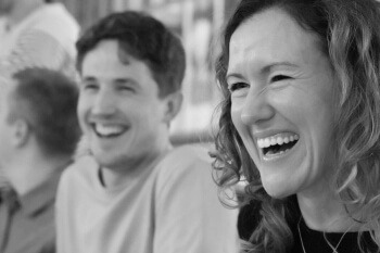 This is a black and white close-up image focusing on the faces of a man and a woman who are both laughing heartily. The woman is in the foreground, with a wide, genuine smile showing her teeth. The man is slightly out of focus behind her, also laughing with his eyes closed or squinting slightly. The image conveys a sense of positive energy, enjoyment, and camaraderie in the working or conference environment.