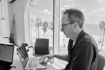 This is a black and white image showing a man working on a laptop at a desk. He is viewed in profile, wearing glasses and a simple dark shirt, focused on the keyboard. The most striking feature of the image is the large window behind him, offering a clear view of a sunny outdoor scene. The background shows a beach or seaside promenade with palm trees, people walking, and the ocean visible in the distance under a partly cloudy sky. This suggests a modern office setting in a coastal location.
