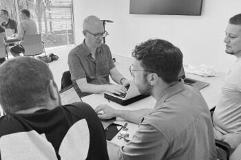 This black and white photo captures a small group of four men engaged in a discussion around a table, likely a team meeting or training session. One man, centrally positioned and wearing glasses, is typing on a laptop and appears to be leading the discussion. The other three men are focused on him or the laptop screen. The overall setting is a modern, indoor space with simple office furniture, emphasizing collaboration and focused work.