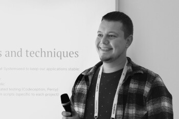 This is a black and white image of a younger man standing and smiling broadly while holding a microphone in his left hand. He is wearing a plaid, possibly flannel, shirt over a patterned t-shirt. He has a lanyard around his neck, indicating he is likely at a conference or event. Behind him is a large white display or banner with text, partially visible, that includes the words "s and techniques" and mentions a company name, "Systemseed," in a context about keeping applications stable. This suggests he is a speaker or presenter.