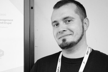 A man with a goatee and short dark hair is smiling directly at the camera. He is wearing a black t-shirt and an event lanyard, standing next to a sign with text partially visible on the left.