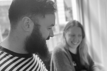 Black and white photo of two people in conversation, smiling. The person in the foreground, wearing a striped shirt and with a full beard, is smiling broadly and looking down. The person in the background is slightly out of focus and is also smiling.