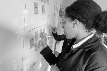 Black and white photo of a person wearing glasses and a dark jacket, writing on a sticky note attached to a whiteboard. Several other sticky notes are visible on the board, suggesting a brainstorming or workshop session. A thin curved line is drawn over some of the notes.