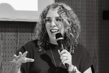 Black and white photo of a person with curly hair and glasses speaking into a microphone. The person is looking forward, their mouth is open as if mid-sentence, and their left hand is raised in an expressive gesture. The background has a vertically paneled wood texture.