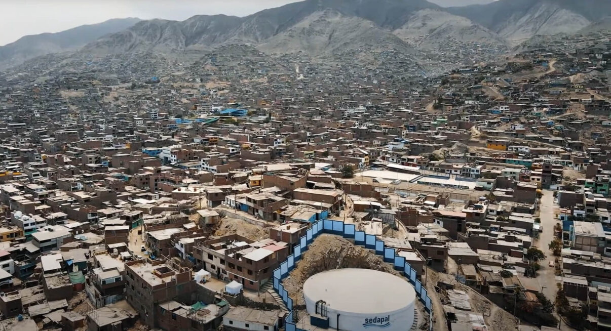 An aerial view of Lima, Peru, featuring dense brick housing sprawling across arid, terraced hillsides under a hazy sky. A large white water tank marked "sedapal" sits prominently in the foreground.