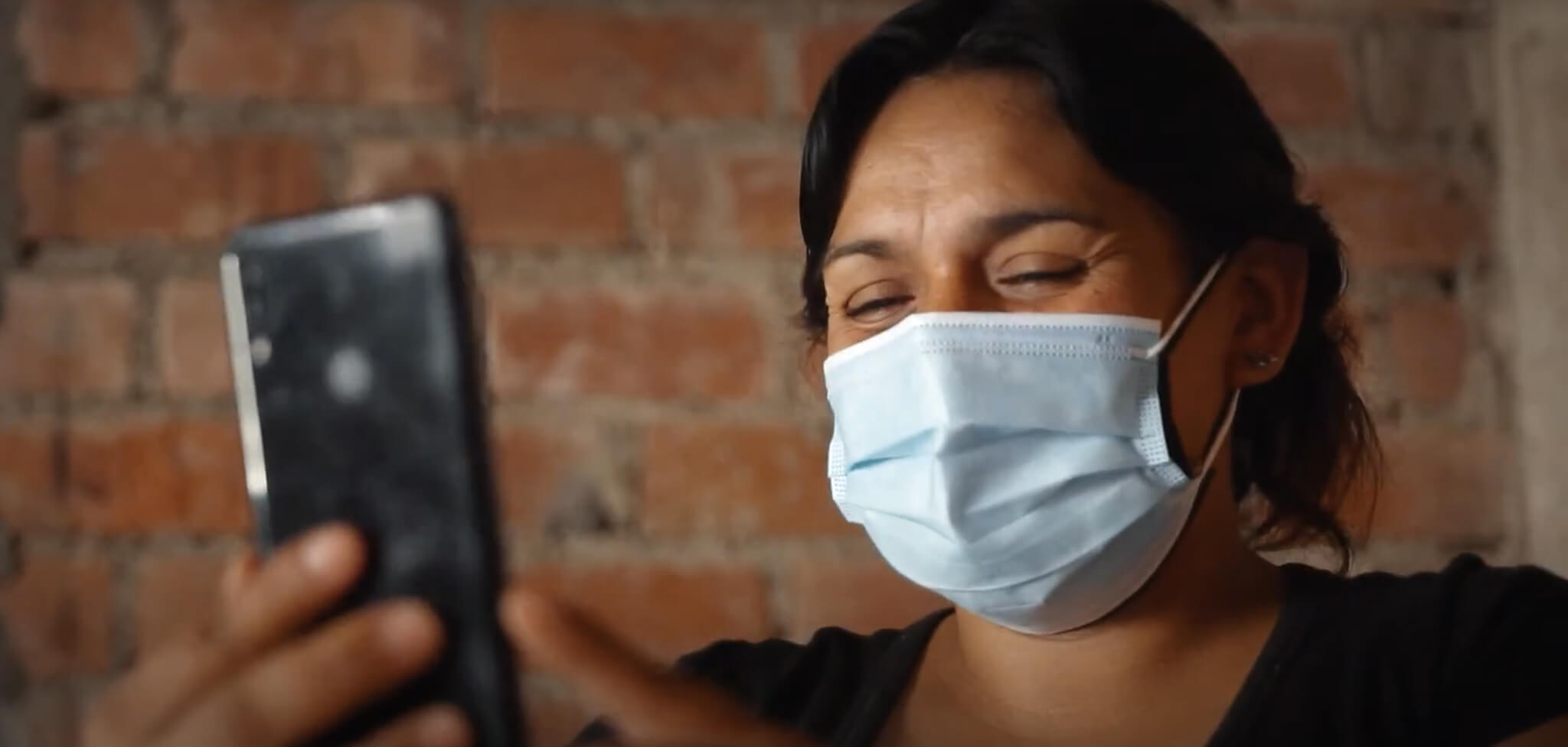 A close-up photograph shows a woman wearing a light blue face mask and black top, holding up a black mobile phone and smiling at the screen, which displays the EQUIP competency tool interface. She is in a building with an exposed brick wall in Peru.