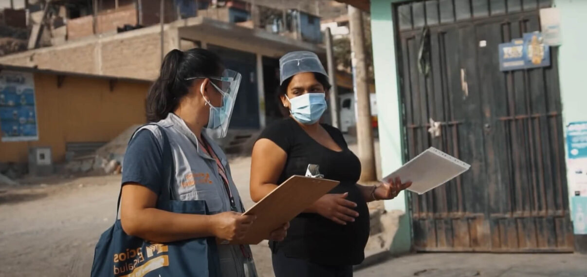 Two women wearing face masks and plastic face shields stand on a dirt road outdoors during the daytime. The woman on the left, wearing a grey vest with a blue bag, holds a wooden clipboard and looks towards the woman on the right. The second woman, who appears pregnant, wears a black t-shirt and holds a clear plastic folder with papers, her hand resting on her stomach. They are in front of a light blue building with a dark metal gate, and in the background are hillside houses.