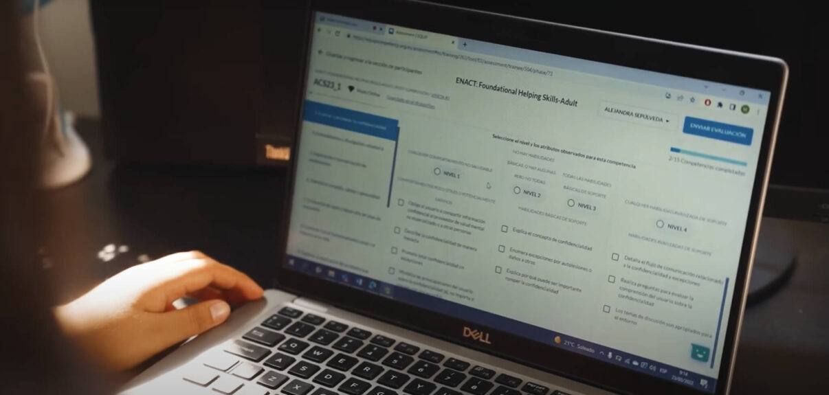 A close-up, over-the-shoulder shot showing a person's hand resting on the keyboard of a Dell laptop. The screen displays a web-based assessment interface titled "ENACT: Foundational Helping Skills-Adult." The content is in Spanish, featuring a form with radio buttons and checkboxes for evaluating specific skills