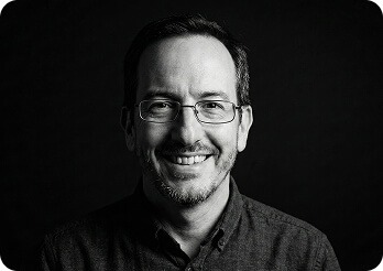 A black and white headshot of Dan Evans smiling warmly, wearing rectangular glasses and a button-down shirt against a dark background.