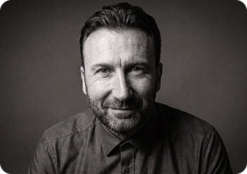 A black-and-white studio portrait of a man with a beard, smiling gently against a solid black background. He is wearing a smart button-up shirt with the collar fastened all the way to the top button, but without a tie. The lighting is dramatic and high-contrast, highlighting his face.