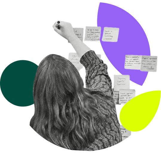A black and white photograph, seen from behind, shows a person with long, wavy hair wearing a patterned sweater, writing on a sticky note with a marker. Their right arm is raised. Numerous handwritten sticky notes are arranged on the surface in front of them. The entire scene is set against a white background with three large, abstract colored shapes: a purple oval, a dark green circle, and a lime green leaf shape.