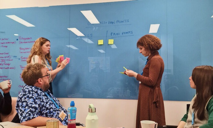 A medium shot of four people in a meeting room gathered around a table. On the left, a man with glasses wearing a blue and white patterned shirt and a woman with curly blonde hair are seated at a light wood table, with a woman standing between them. The woman standing, who has wavy blonde hair and is wearing a white shirt, is adding pink sticky notes to a blue glass whiteboard. The man is looking up at her. To the right of the standing woman is another woman with short, curly brown hair wearing a long-sleeved brown polka dot dress, who is looking at yellow sticky notes in her hands. Further right, another woman with long, straight brown hair wearing a green sweater vest is seated, looking towards the whiteboard. The blue whiteboard has the words "GOALS + PAIN POINTS" written at the top, with "GOALS" on the left and "PAIN POINTS" on the right. There are several yellow and pink sticky notes on the board. On the table, there are mugs, a water bottle, and a metal canister. The background shows a glimpse of a window and office cubicles.