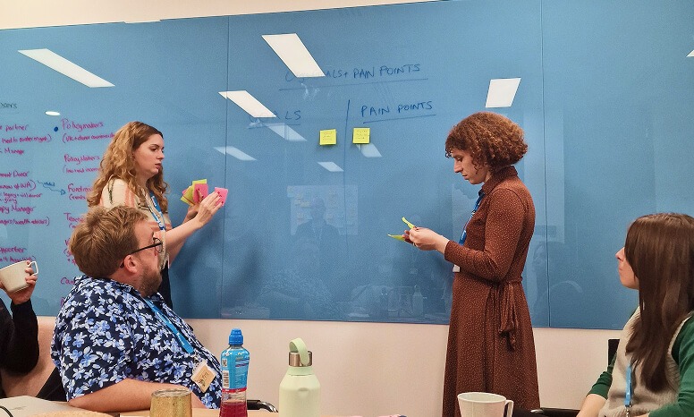 A medium shot of four people in a meeting room gathered around a table. On the left, a man with glasses wearing a blue and white patterned shirt and a woman with curly blonde hair are seated at a light wood table, with a woman standing between them. The woman standing, who has wavy blonde hair and is wearing a white shirt, is adding pink sticky notes to a blue glass whiteboard. The man is looking up at her. To the right of the standing woman is another woman with short, curly brown hair wearing a long-sleeved brown polka dot dress, who is looking at yellow sticky notes in her hands. Further right, another woman with long, straight brown hair wearing a green sweater vest is seated, looking towards the whiteboard. The blue whiteboard has the words "GOALS + PAIN POINTS" written at the top, with "GOALS" on the left and "PAIN POINTS" on the right. There are several yellow and pink sticky notes on the board. On the table, there are mugs, a water bottle, and a metal canister. The background shows a glimpse of a window and office cubicles.