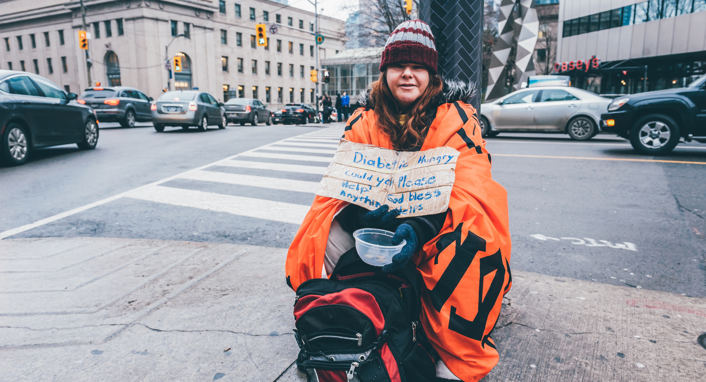 Homeless person on street corner wearing a hat and holding a sign