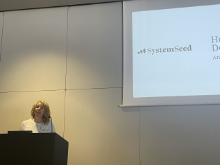 Elise West stands at a podium, in the background is a large presentation screen showing the SystemSeed logo